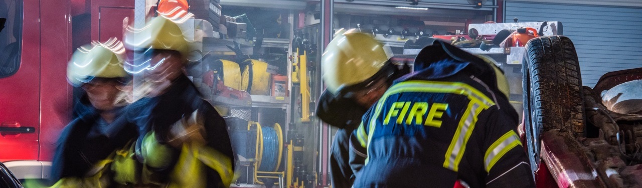 Close up firefighters breaking the door on upside down car, fire engine in background.